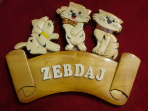 Houten bordje met daarop de naam Zebdaj. Het bordje lijkt op een lint met opgekrulde uiteinden. Bovenop zitten drie vrolijke katjes.